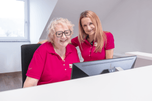 Zwei Mitarbeiterinnen des Mammographie Screening Niederbayern in einheitlichen magentafarbenen Shirts lächeln und schauen gemeinsam auf einen Computerbildschirm in einem hellen Untersuchungsraum – Symbol für kompetente, freundliche Betreuung und die hochwertige Brustkrebsfrüherkennung im regionalen Screening-Programm für Frauen von 50 bis 75 Jahren.