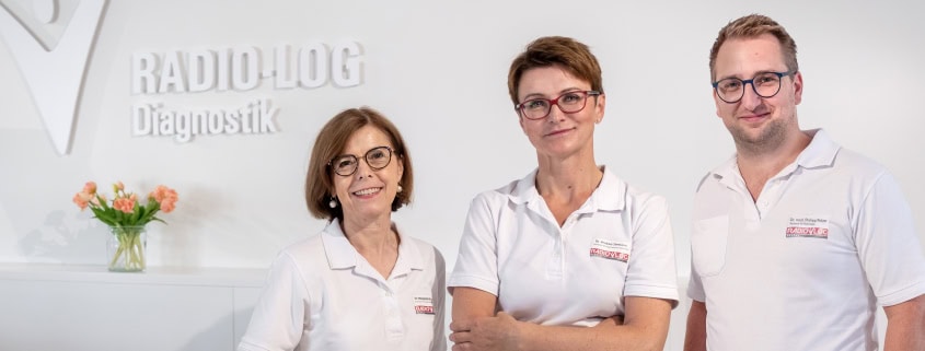 Drei medizinische Fachkräfte in weißen Kitteln stehen am Empfangstresen vor einem Schild mit der Aufschrift "RADIO-LOG Diagnostik" und setzen sich engagiert für das Mammographie-Screening Niederbayern zur Früherkennung von Brustkrebs ein. Ein Blumenstrauß auf dem Tresen sorgt für eine freundliche Atmosphäre im zertifizierten Programm zur Brustkrebsvorsorge für Frauen von 50 bis 75 Jahren.