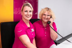 Zwei Mitarbeiterinnen in rosa Hemden am Empfang von Mammographie Screening Niederbayern, eine lächelt, eine telefoniert.