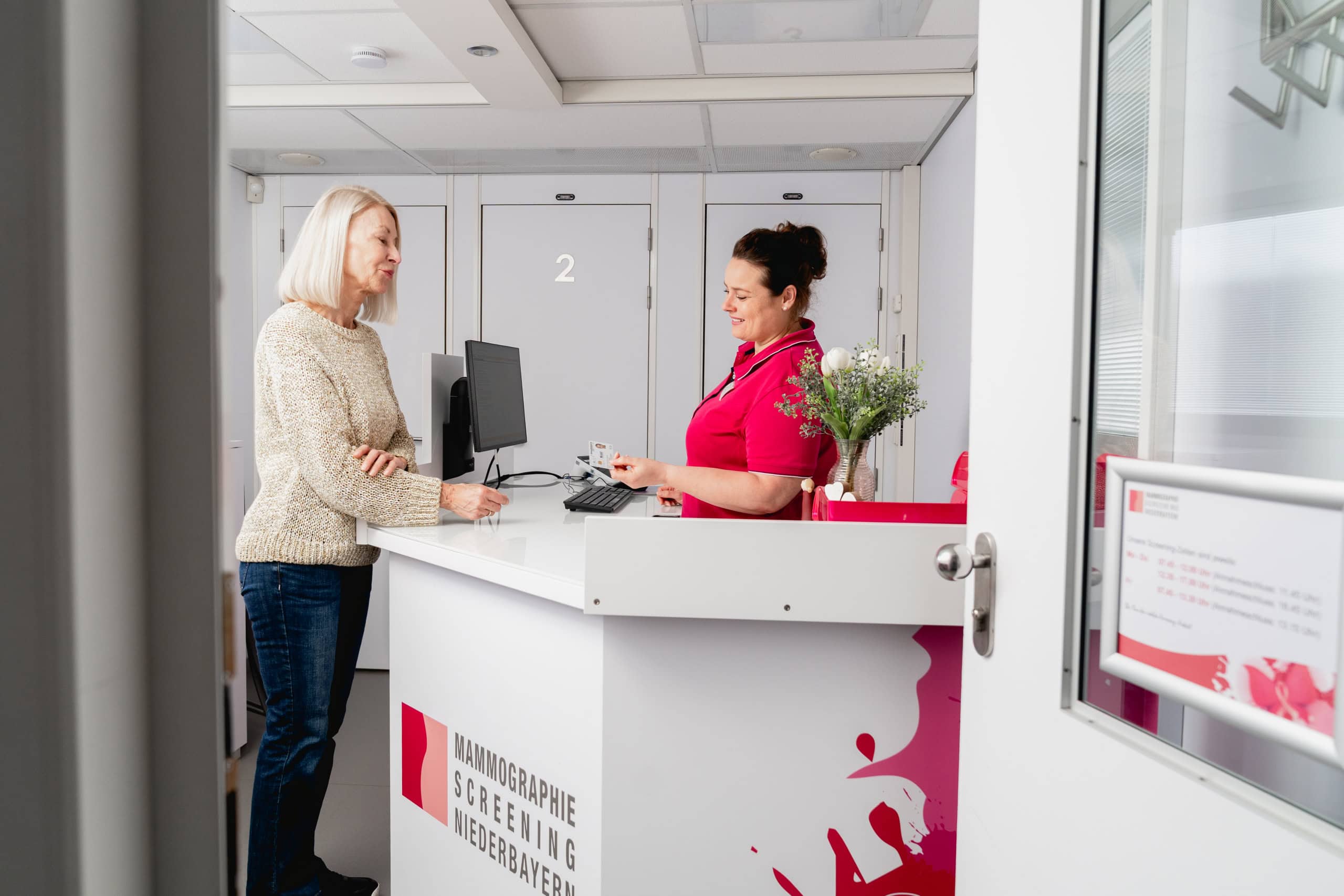 Eine Frau spricht am Empfang eines Mammographie-Screening-Zentrums in Niederbayern mit einer Mitarbeiterin in rosa Kittel.