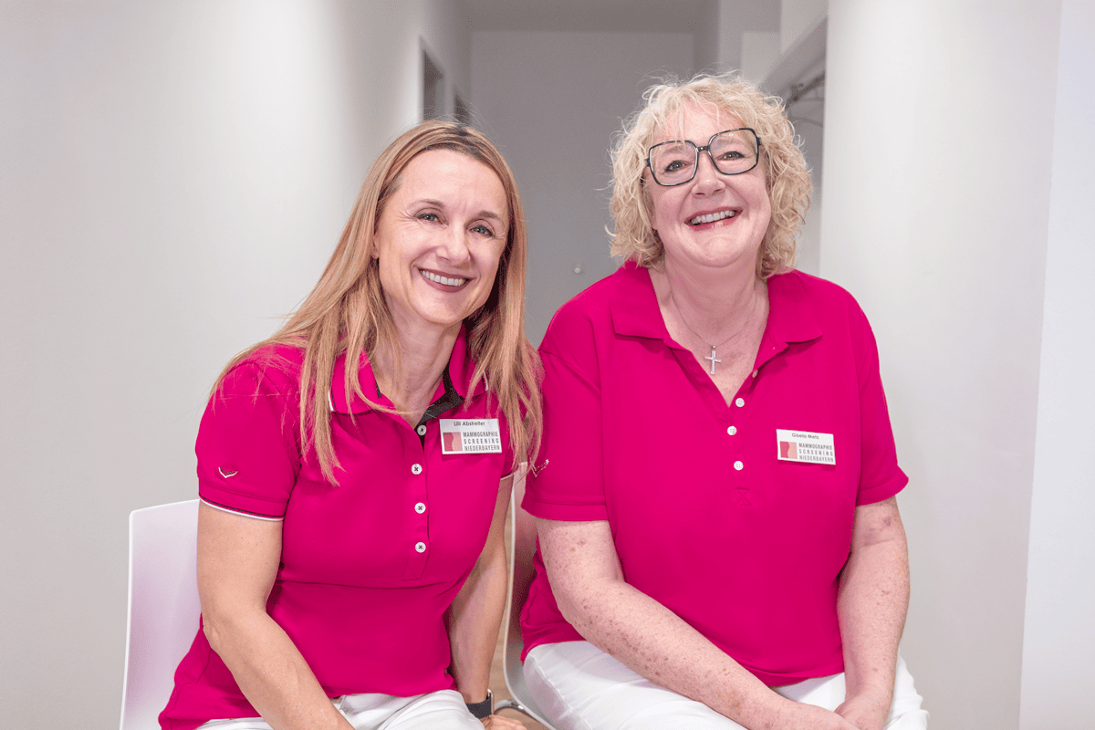 Zwei Frauen in rosa Poloshirts mit Namensschildern, die freundlich lächeln, im Beratungsbereich des Mammographie-Screenings Niederbayern.
